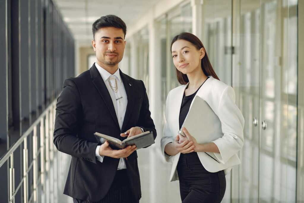 Confident well dressed multiracial coworkers standing in modern office corridor while holding laptop and notebook in hands and looking at camera