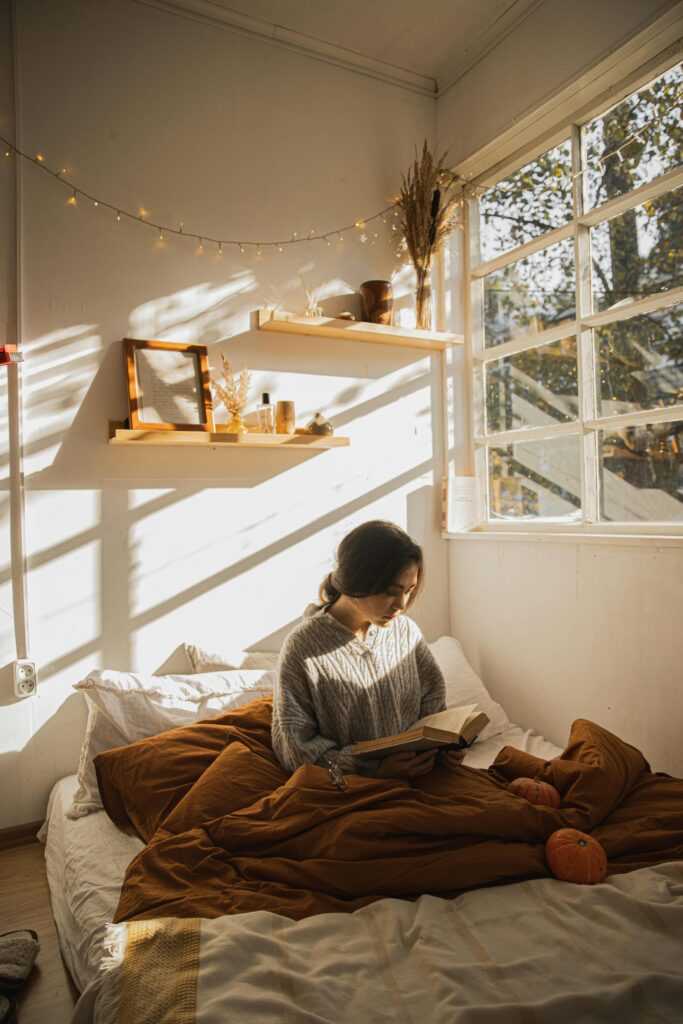 Woman reading in a sunlit bedroom, surrounded by autumn decor and pumpkins.