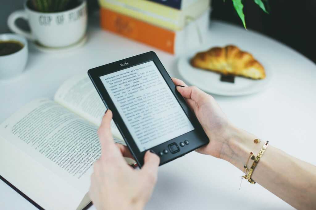 A woman holds a Kindle while reading with a book and croissant on a table.