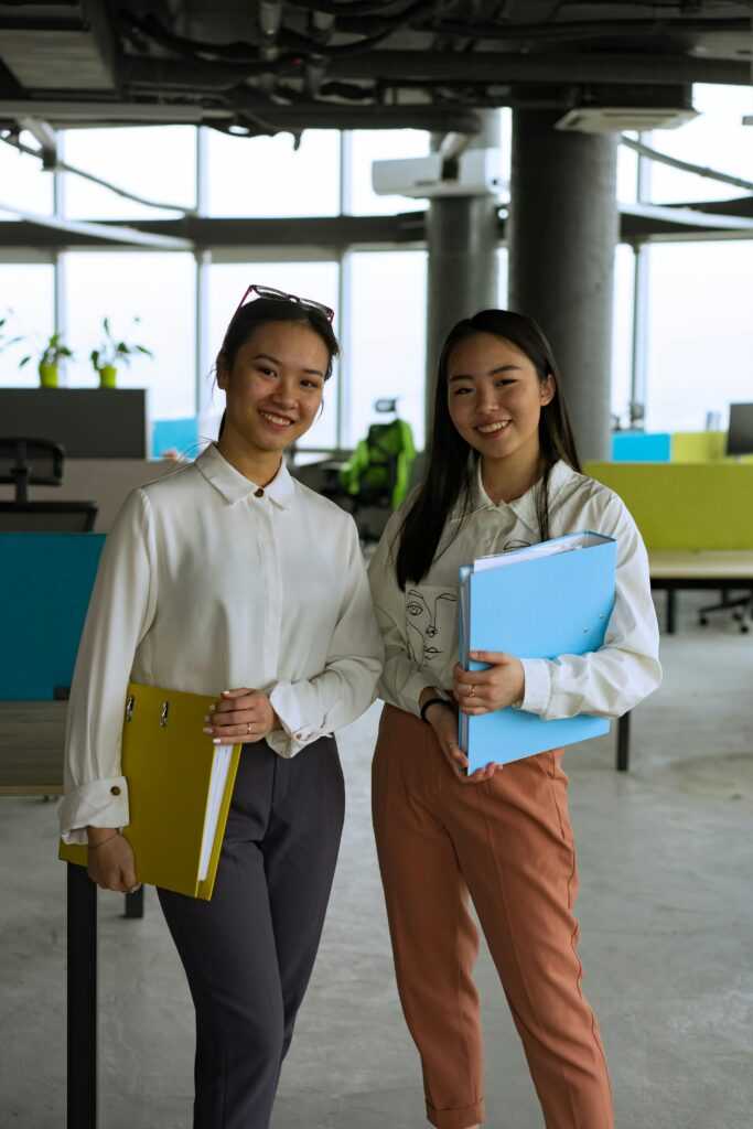 Two smiling women in a modern office holding binders, exuding professionalism and positivity.